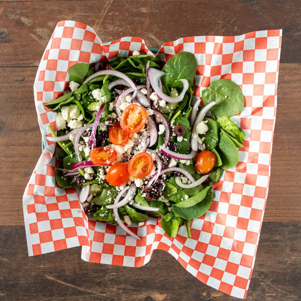 Spinach salad with tomatoes, red onions, olives, and feta cheese in a checkered paper-lined container.