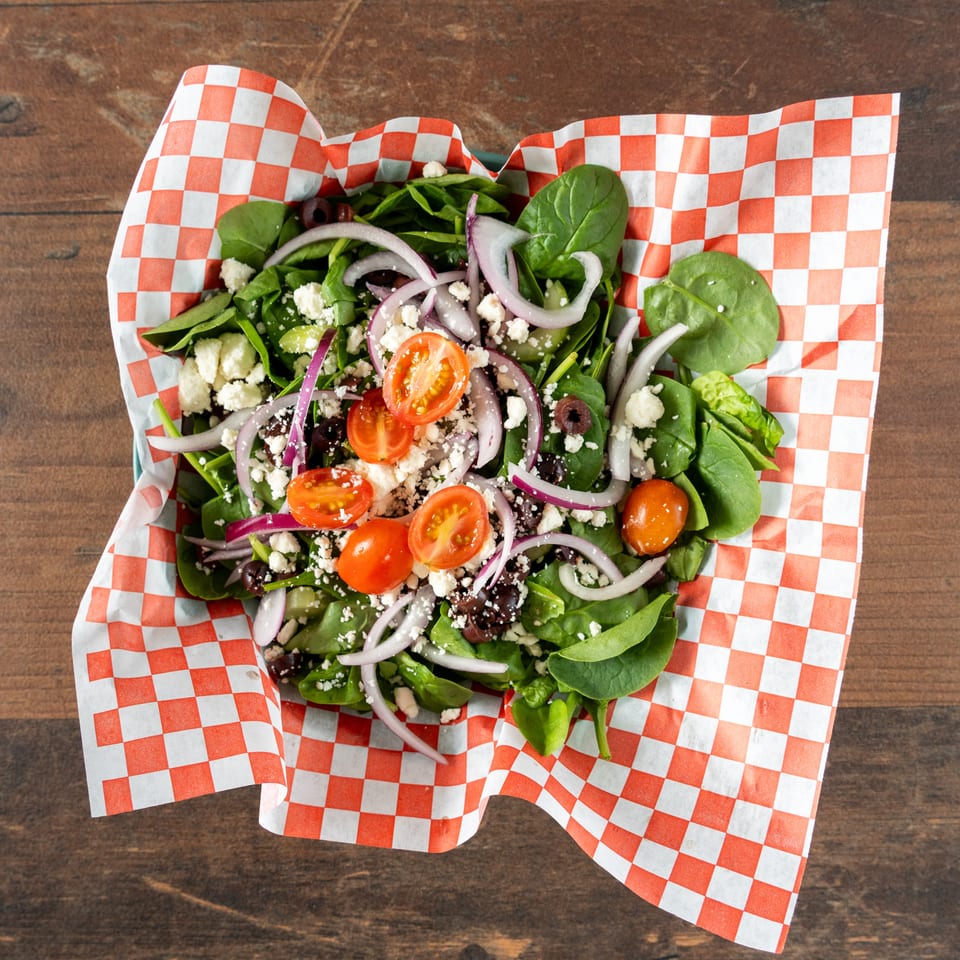 Spinach salad with tomatoes, red onions, olives, and feta cheese in a checkered paper-lined container.