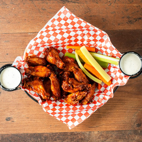 Basket of sauced chicken wings with celery, carrots, and two dipping sauces on a wooden table.