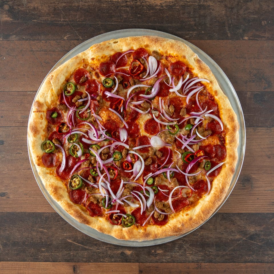 Whole pizza with pepperoni, sausage, red onions, and jalapeños on a wooden table.